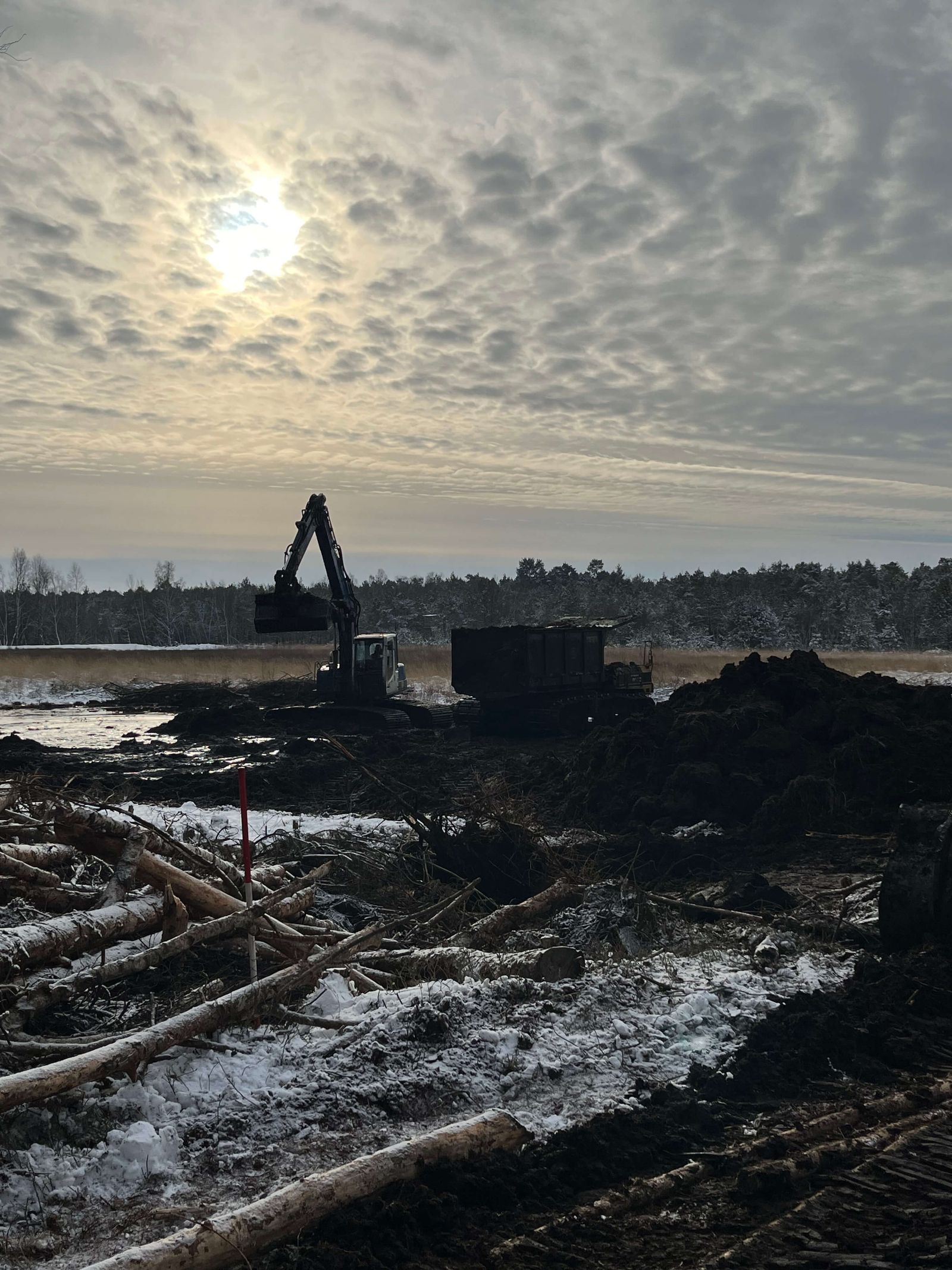 Bagger arbeitet auf verschneitem, matschigem Gelände unter bewölktem Himmel in winterlicher Landschaft.